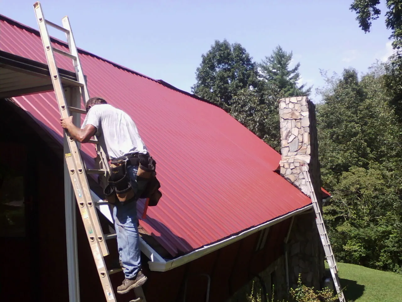 Roofer on a ladder beside a red metal roof and stone chimney — Soffit & Fascia Repair by Roofing Brothers in Gerber